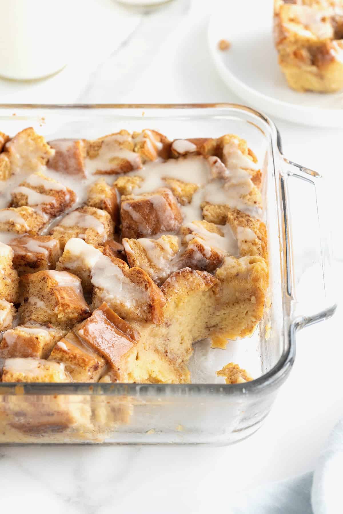 A square glass baking dish of bread pudding with white icing drizzled on top. The bottom right hand corner of the dish has the bread pudding scooped out.